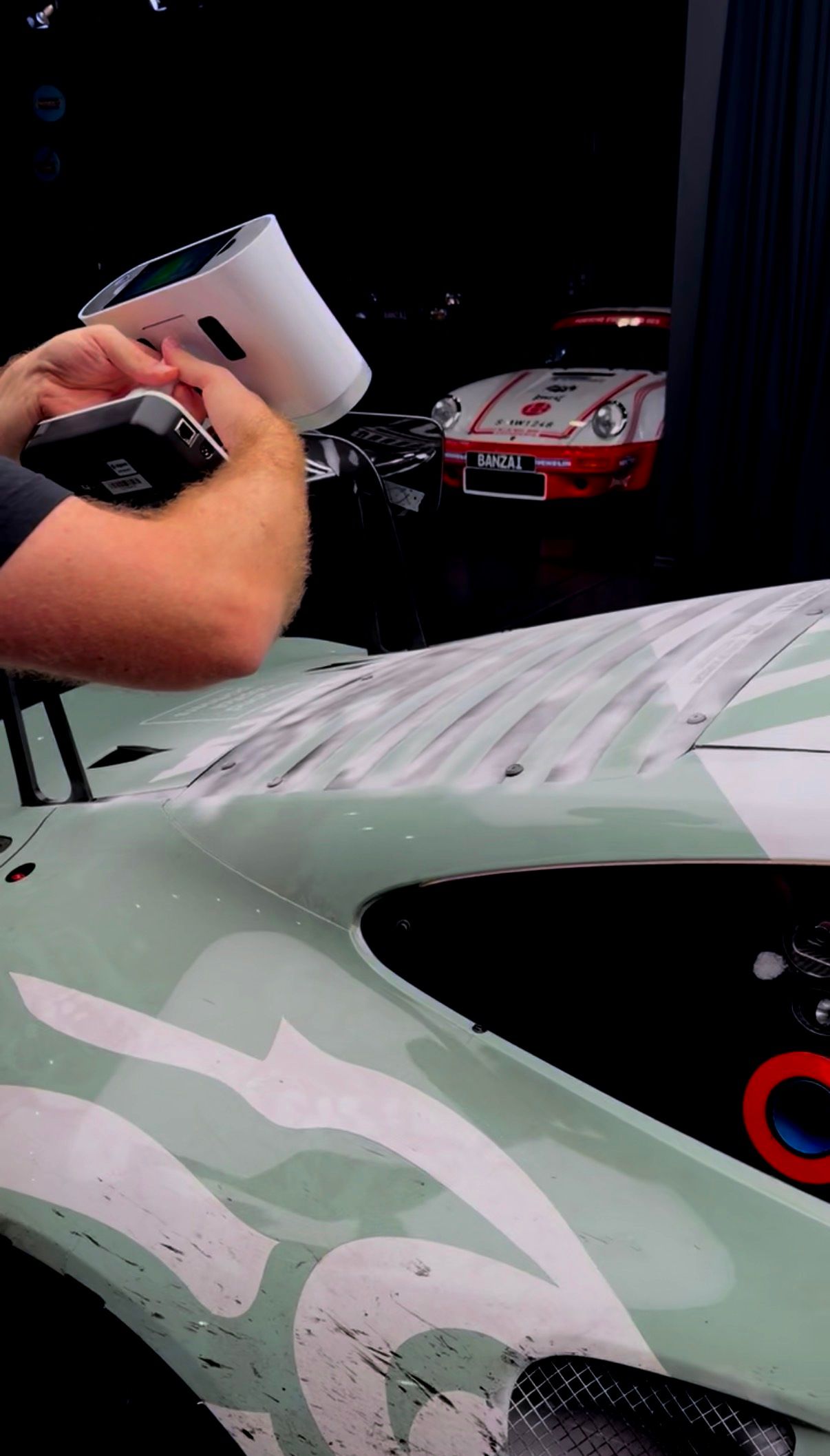 Hand-held 3D scanner capturing the hood of a Porsche GT3 race car in the Banzai Legacy Lab workshop, a red vintage 911 visible in the background