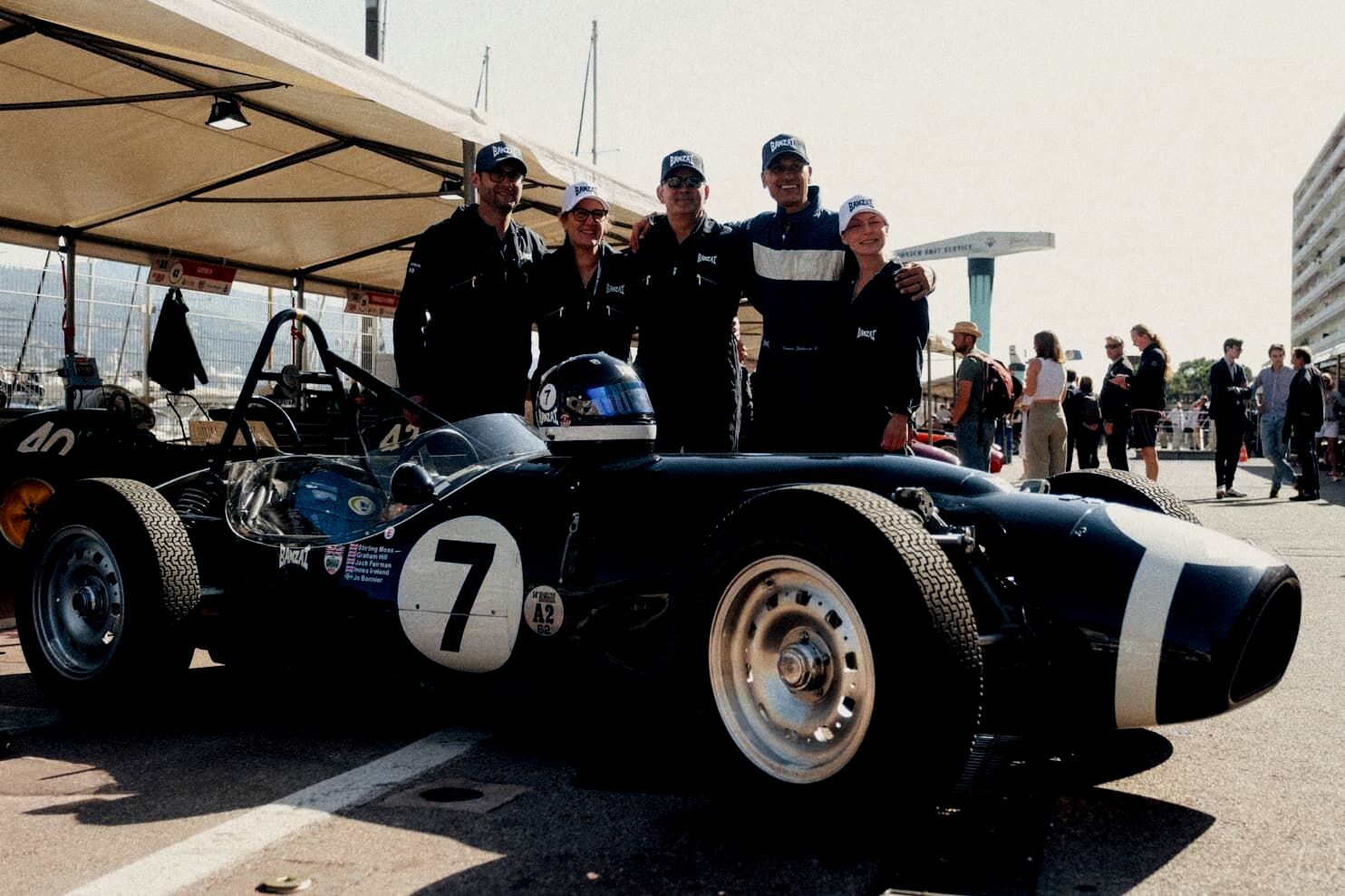Team Banzai Legacy Lab crew posing together beside a vintage open-wheel racer number 7 in the pit lane at a classic race meeting